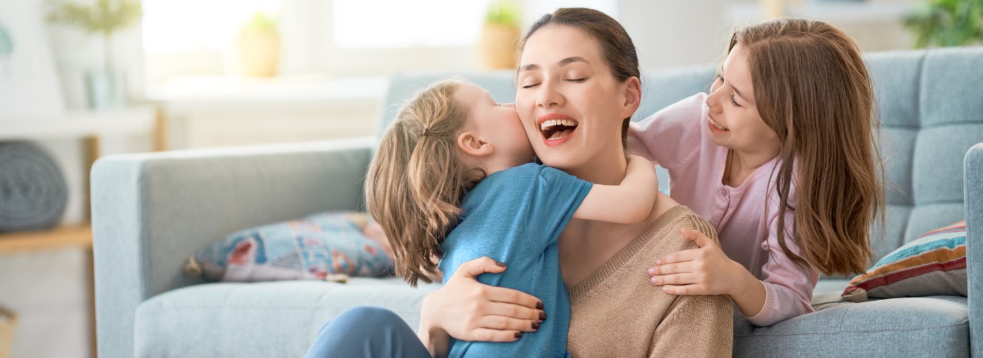 Een moeder zit lachend en ontspannen op de grond met haar twee kinderen, die haar liefdevol omhelzen in een gezellige, lichte woonkamer.