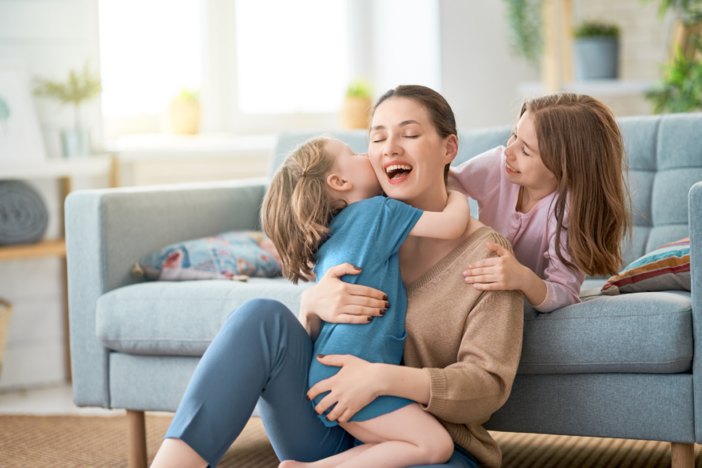 Een moeder zit lachend en ontspannen op de grond met haar twee kinderen, die haar liefdevol omhelzen in een gezellige, lichte woonkamer.