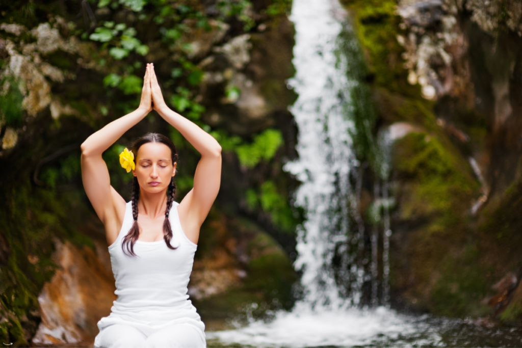 Vrouw met gesloten ogen zit voor een waterval, met handen boven haar hoofd en handpalmen tegen elkaar, die vrouwelijke energie, balans en innerlijke rust symboliseert.