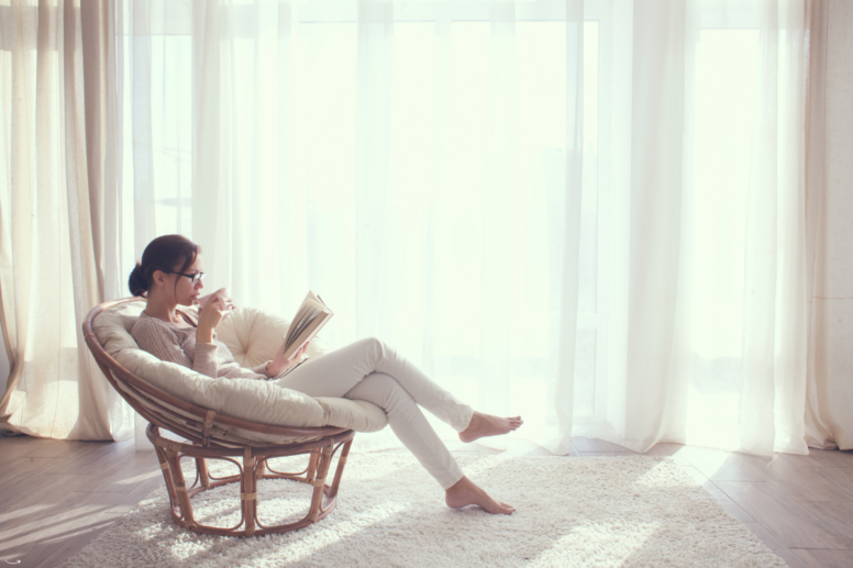 en vrouw die ontspannen in een stoel zit, een boek leest en een slok thee neemt, symboliseert de rust en zelfzorg die hoogsensitieve personen waarderen.