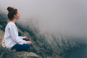 Een vrouw zit in meditatiehouding op een rotsachtig bergpad met een mistig uitzicht op de bergen en de zee.