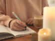 en vrouw schrijft in een journal met een turquoise pen terwijl ze geniet van een kop koffie. Kaarsen branden op de voorgrond, wat een rustgevende sfeer creëert.