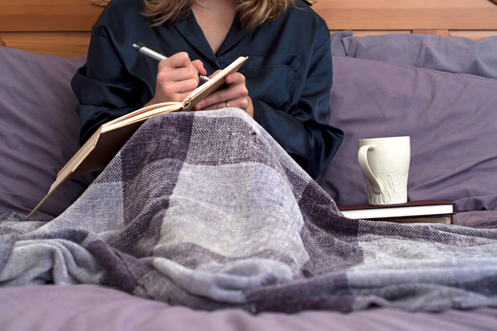 Vrouw schrijft, zittend in bed met een deken over zich heen, in haar journal met een beker thee naast zich.

