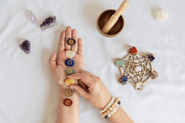 Foto ter illustratie van de online Chakra Healing training: links een hand met zeven chakra-edelstenen, rechts edelstenen op een houten symbool en een klankschaal op de achtergrond. De afbeelding symboliseert het balanceren van energie en het ervaren van diepe heling tijdens de cursus.