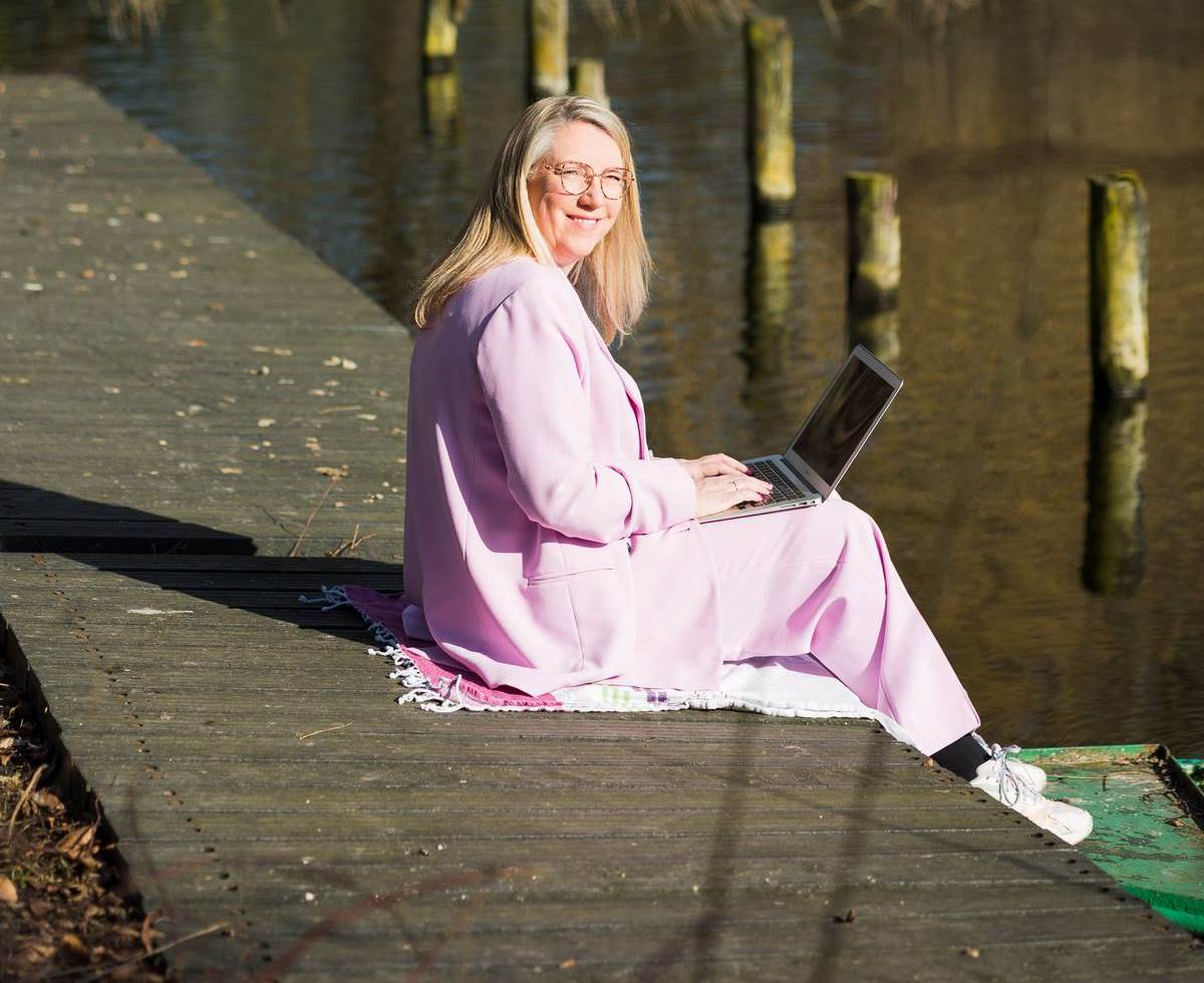 Sandy Vivaldi, holistisch therapeut, zittend aan de rand van het water met haar laptop op schoot.