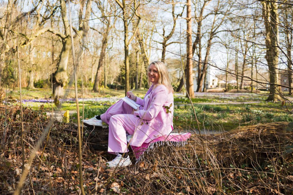 Sandy Vivaldi, holistisch therapeut zit ontspannen op een boomstam in het bos te schrijven in haar journal.