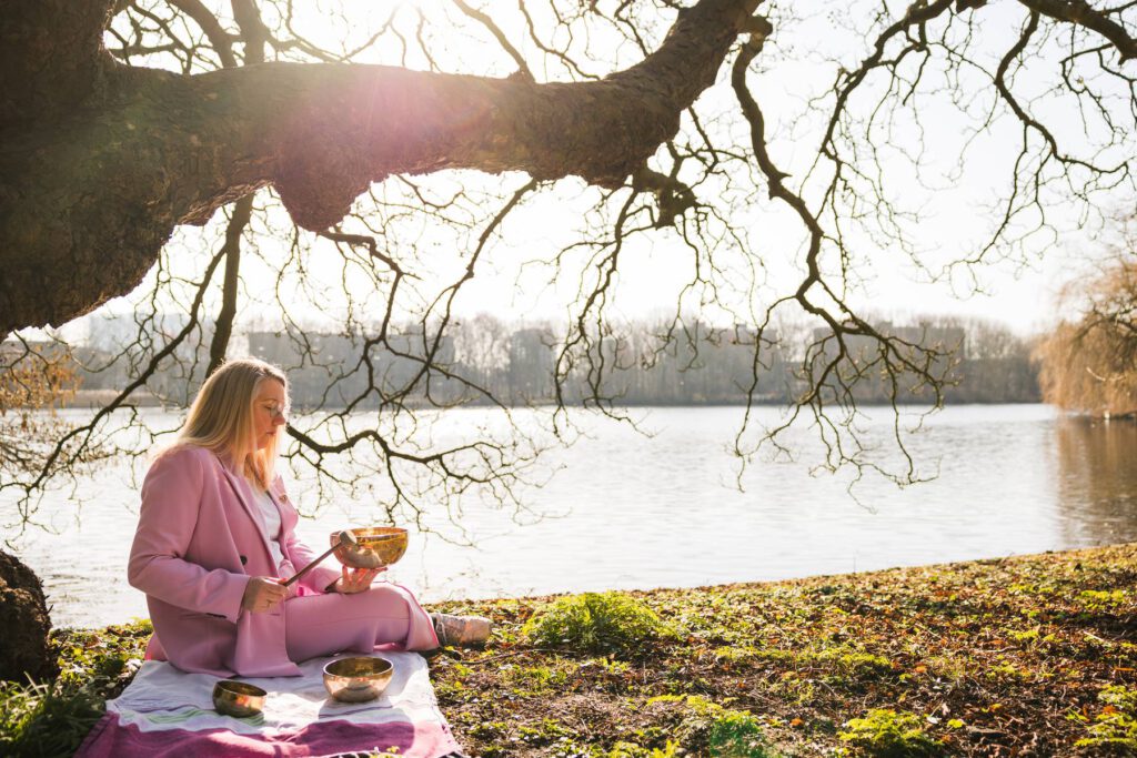 Sandy Vivaldi, holistisch therapeut zit in het bos, aan het water onder een boom en bespeelt een klankschaal.